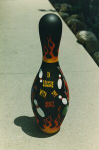 Bowling pin painted with the text "In League With The Devil" amongst flying bowling pins and hotrod style flames.